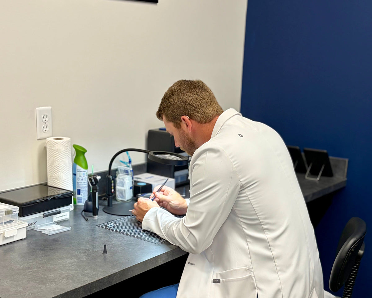 A man in a white doctor's coat and blue pants sits at a desk in a clinic, writing in a notebook. The room has a dark gray desk, a black office chair, a blue accent wall, and medical supplies on the counter.